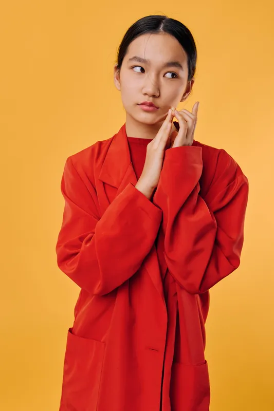 Young woman wearing bright red coat standing against yellow background looking sideways with thoughtful expression and hands touching face in casual fashion portrait.
