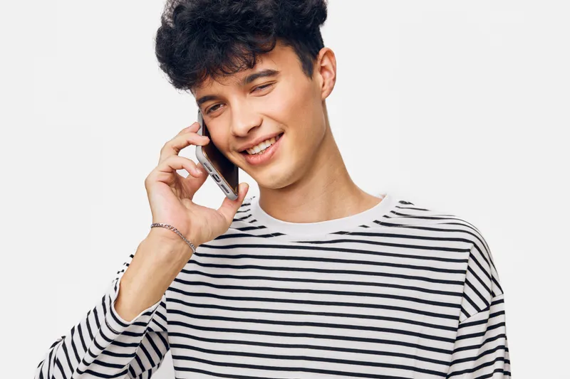 smiling young man talking on smartphone wearing casual striped shirt with modern hairstyle and light background, expressing happiness and friendliness