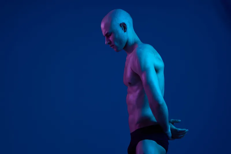 A contemplative man in a unique light, showcasing his expression against a striking blue background. The image features a thoughtful pose and minimal clothing.