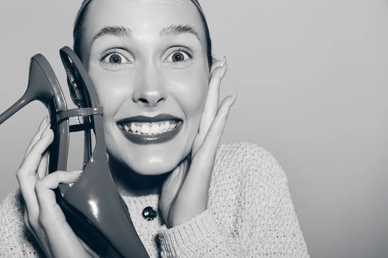 Young white woman with light skin smiles excitedly holding red high heel shoes near face in studio. Concept of fashion, happiness, and footwear.