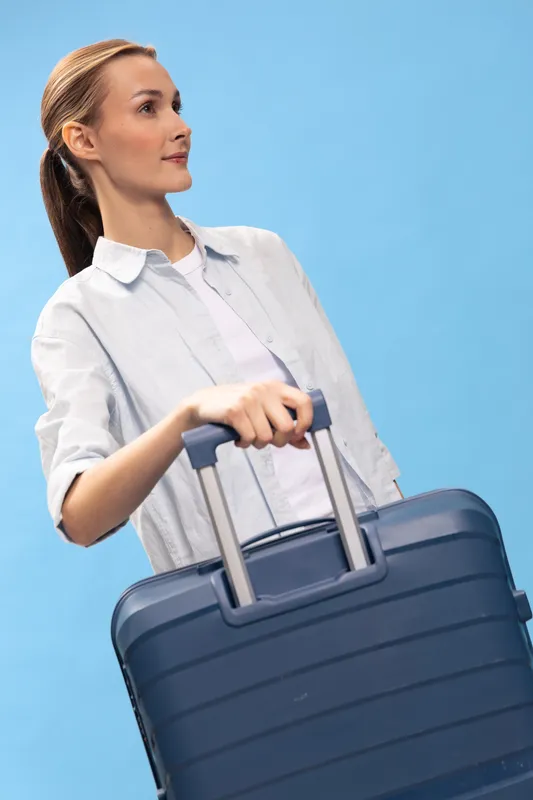 Young woman with suitcase looks sideways on blue background. Travel concept, casual outfit, luggage, and light mood for vacation or business trip.