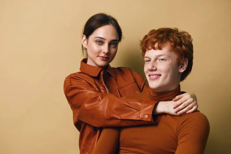 Young woman and man with brown hair wearing brown outfits posing together for portrait with warm expressions on beige background in casual modern fashion style.