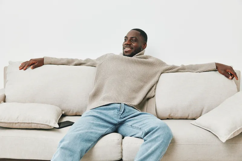 Relaxed young Black man enjoying leisure time on a beige couch, wearing a cozy sweater and jeans, exudes happiness in a modern living room setting