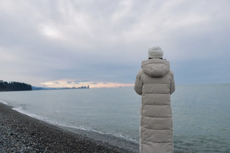 Solitude, coastline, winter, contemplation, seascape, person in a long coat standing on a beach, gazing at the calm horizon under overcast light.