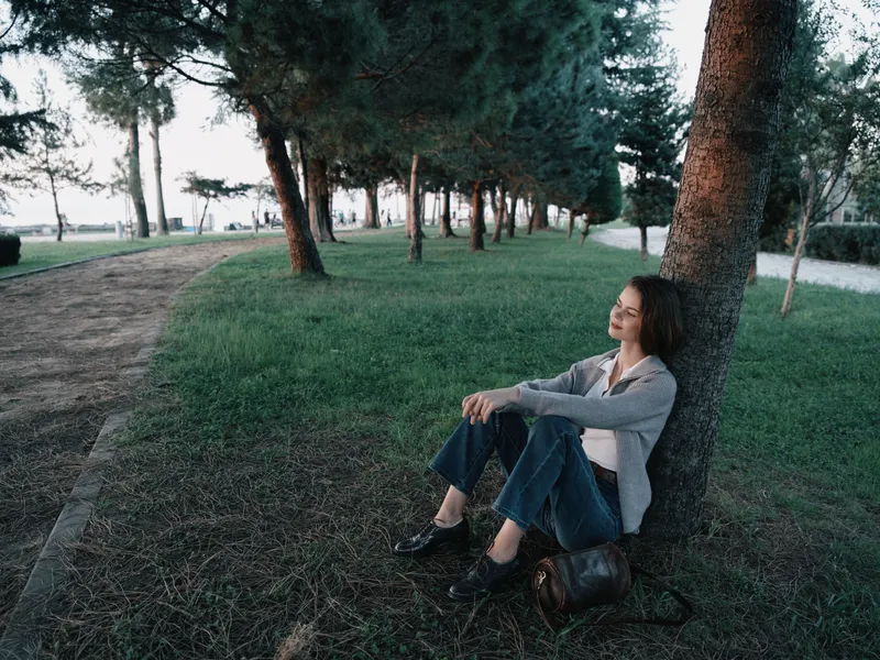 A thoughtful young woman sits against a tree in a park, relaxed and contemplative, with soft natural light creating a tranquil outdoor atmosphere.