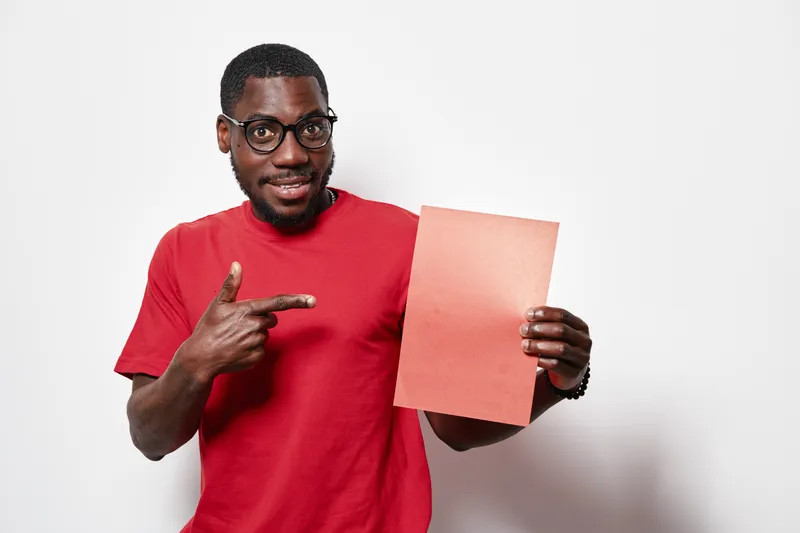 African American man with dark skin and glasses wearing red shirt pointing at blank paper for presentation or advertisement on white background. Smiling, confident and casual pose.