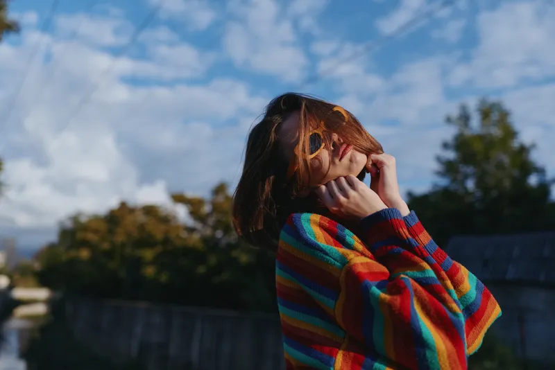 Young woman in her early twenties wearing a colorful striped sweater, outdoor portrait by river, smiling with windblown hair, hands near face, relaxed lifestyle and travel vibe.