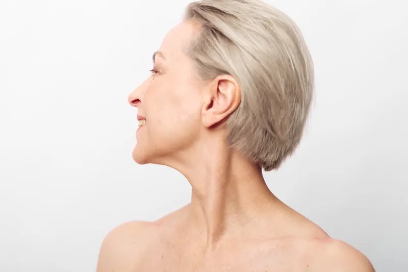 Profile of smiling middle aged woman with short blonde hair and bare shoulders on white background. Concept for beauty, aging, skin care, wellness, and natural look.