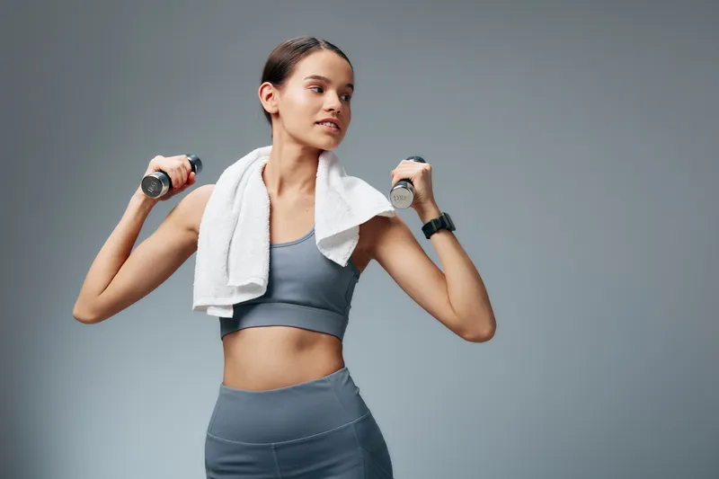 Fit young woman in grey sportswear holding dumbbells with towel draped over her shoulders, standing against a neutral studio background in a confident pose highlighting a toned midriff.