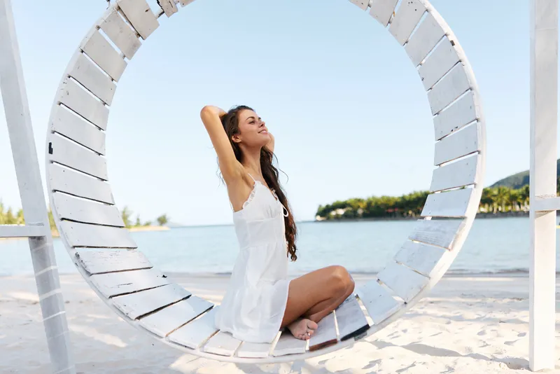 A woman sits in a large circular swing by the seaside, wearing a white dress. Sunlit, peaceful scene with calm expression on a breezy beach.