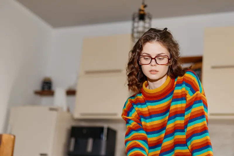 Young woman with curly hair and glasses leans over a kitchen counter in a bright, cozy home setting, wearing a vibrant striped sweater as she concentrates on a task.
