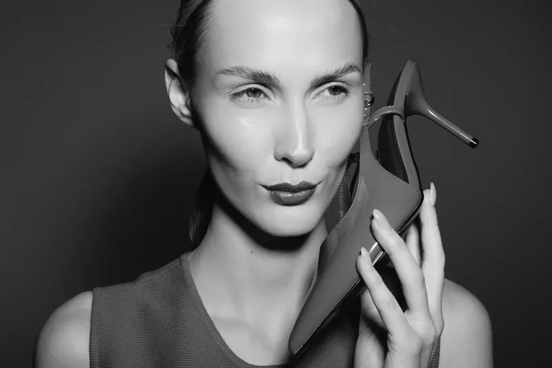 Elegant white woman with fair skin holds high heel shoe near face, posing in studio. Concept for fashion, beauty, and footwear.