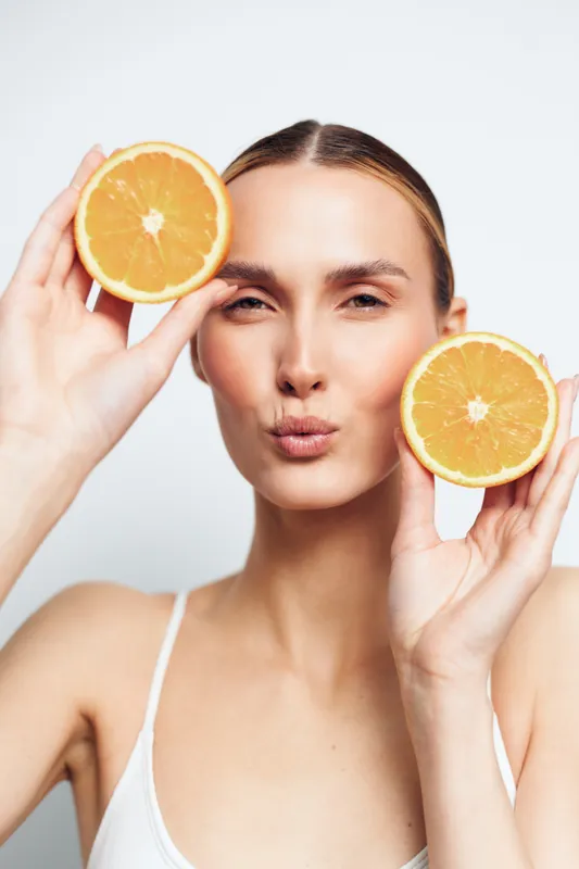 Young woman with clear skin holding two halves of fresh orange fruit near her face. Natural beauty concept with healthy lifestyle and vitamin C. Bright studio lighting, white background.