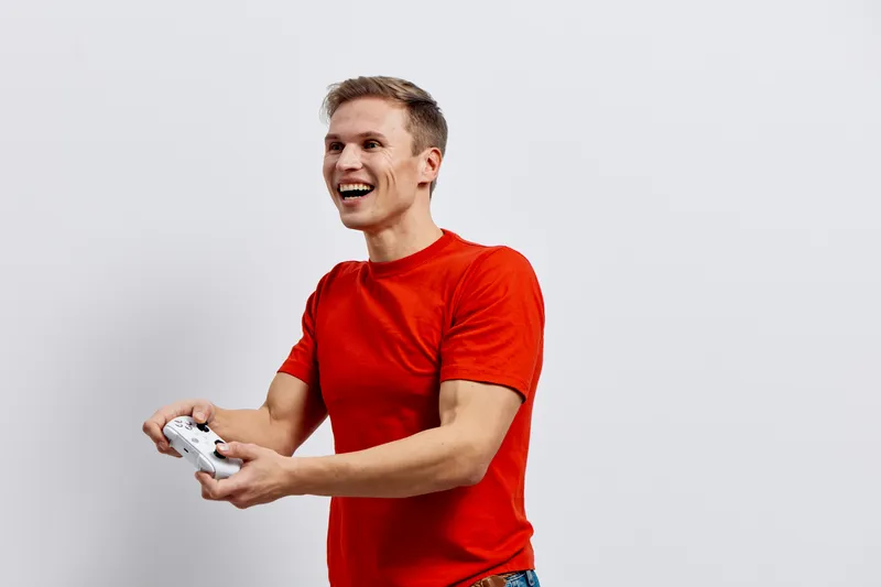 Smiling man with short hair wearing casual red t-shirt holding video game controller isolated on plain white background studio portrait. People lifestyle concept
