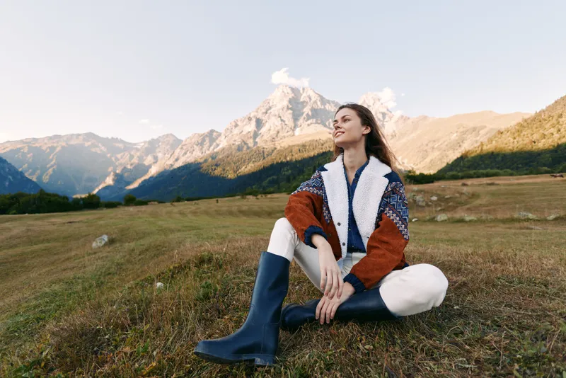 Woman sitting on meadow in front of mountain landscape, outdoor portrait of young woman wearing jacket and boots, relaxed pose in nature with scenic horizon and clear sky.