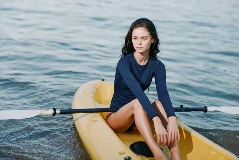 A female rower sits in a yellow single scull on calm water, wearing a dark blue long sleeve top, poised with oar ready as gentle waves glisten nearby.
