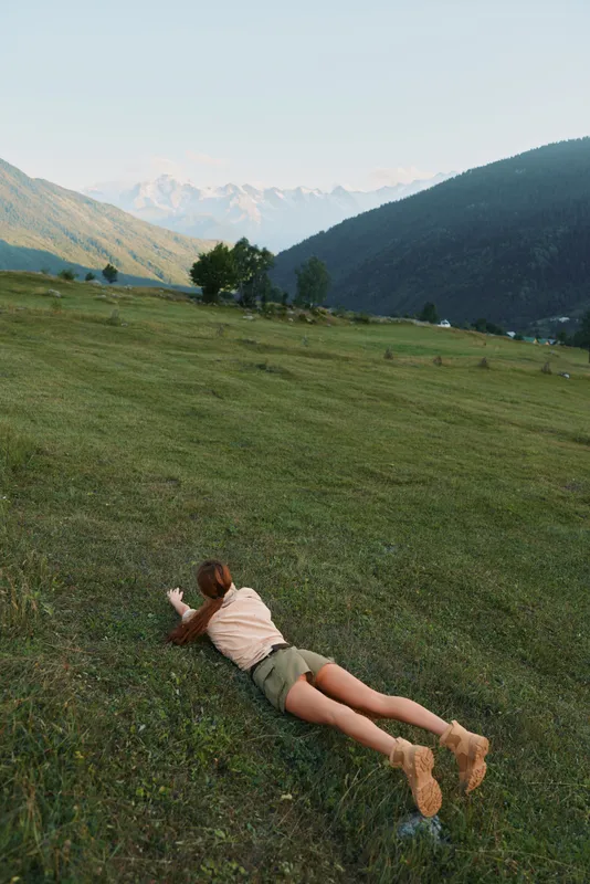 A young woman lies prone on a sunlit grassy hillside in a quiet valley, with distant mountains in view, conveying relaxation, freedom, and connection with nature.