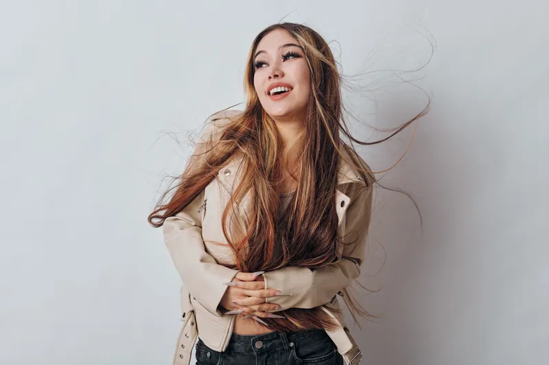 Young woman with long hair smiling and looking away while wearing light jacket and dark pants standing against light background in studio with hair movement.