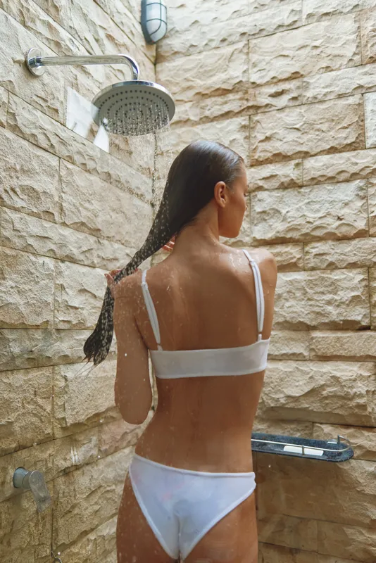 Woman showering outdoors in white underwear with wet braided hair, natural stone wall background, relaxing moment under rain showerhead, wellness and freshness.