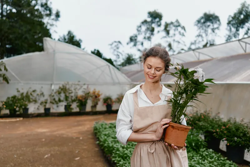 Young woman in a greenhouse holding a potted plant, wearing an apron and smiling, showcasing gardening and nature themes in a vibrant green setting