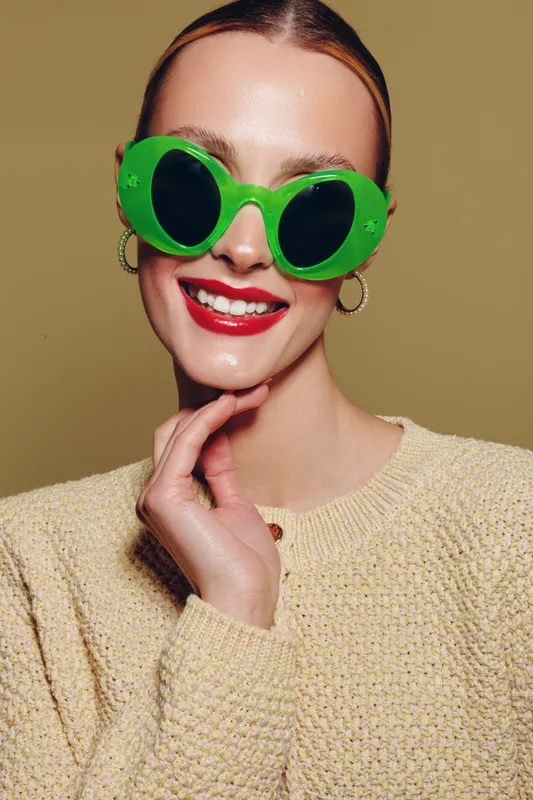 Smiling woman with green sunglasses and red lipstick poses with hand near face wearing beige sweater. Studio portrait with warm background. Concept of fashion, style, and beauty.