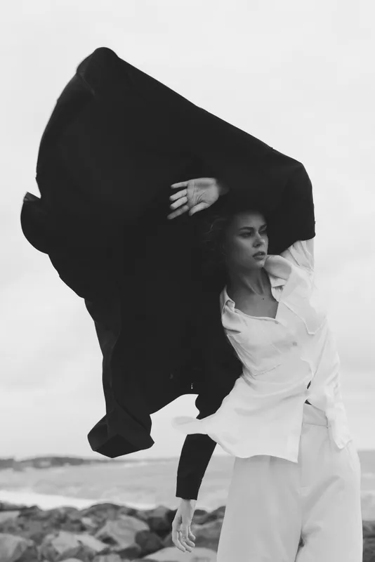 Fashion movement on a windswept beach, a model draped in a flowing coat against the sea, captured in monochrome for a bold, avant garde outdoor fashion moment