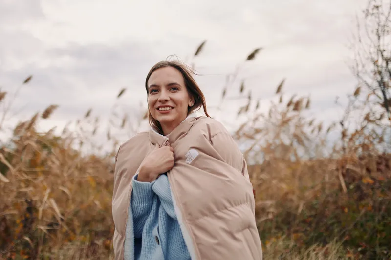 Genuine outdoor portrait of a smiling young woman wrapped in a beige coat and blue knit sweater, conveying authentic emotion and credible everyday lifestyle in tall autumn grass.