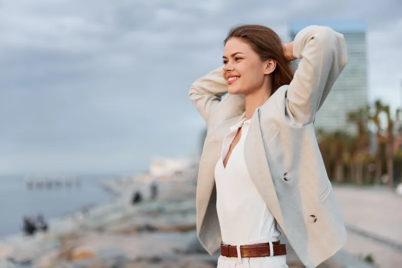 Confident businesswoman enjoying the seaside view, dressed in a light blazer and smiling, with a blurred urban background, showcasing a bright and optimistic mood.