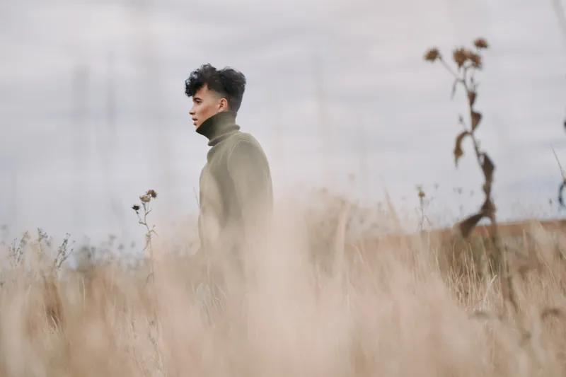 A young man in a turtleneck stands in a windswept field, captured with authentic, documentary style and genuine presence, conveying a realistic, contemplative portrait vibe. (165 characters)