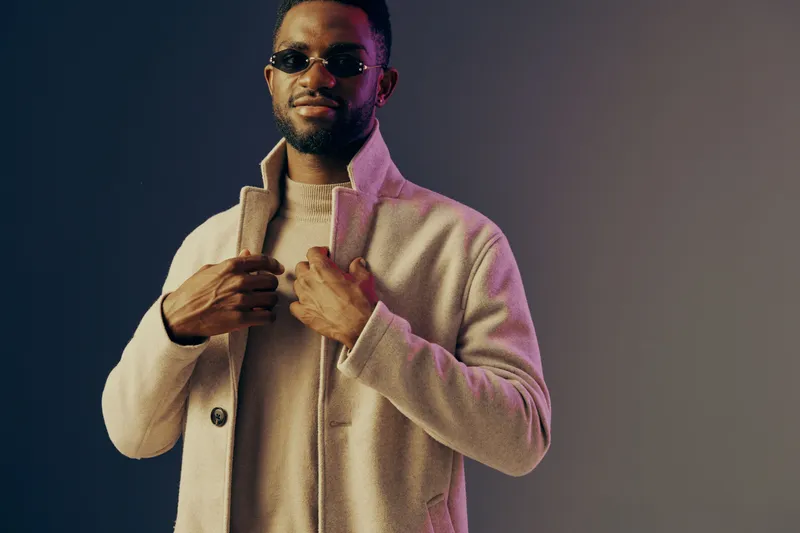 Stylish African American man in a fashionable beige coat posing confidently against a vibrant gradient background, showcasing modern urban fashion