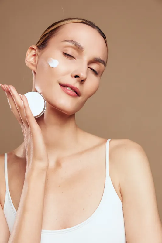 Young woman with clean skin applies face cream on cheek holding cosmetic jar with closed eyes and relaxed expression isolated on beige background in skincare concept.