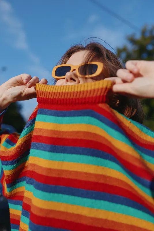 Woman in her mid-20s wearing a colorful striped sweater and yellow sunglasses pulls the collar up playfully outdoors, casual lifestyle portrait under sunny blue sky with joyful mood.