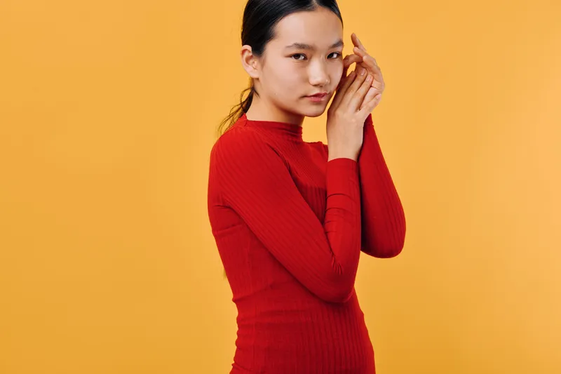 Young woman in red long sleeve shirt standing against yellow background looking at camera with thoughtful expression and natural makeup for portrait photography.