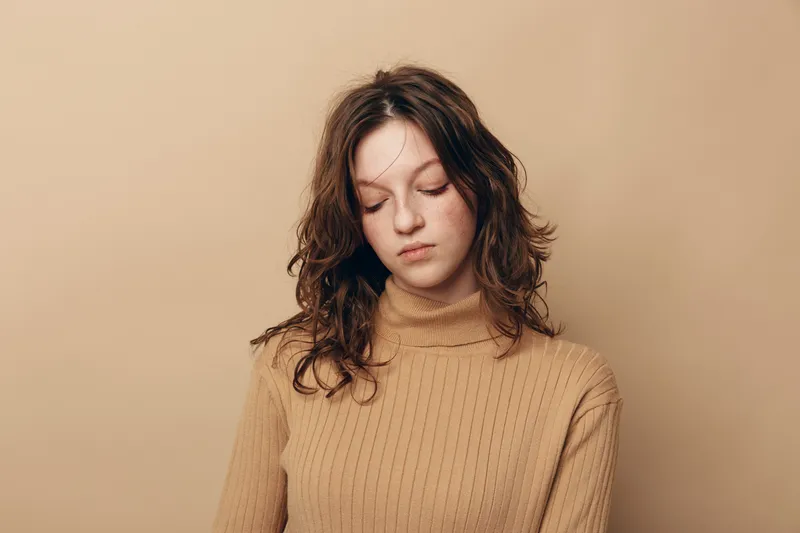 Sad young woman with curly hair wearing beige sweater looks down with closed eyes. Studio portrait on neutral background. Concept of loneliness, sadness, and introspection.