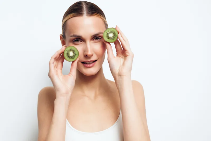 Young woman holding two halves of fresh kiwi fruit near her face, looking at camera, clear skin, minimal makeup, white background, healthy lifestyle concept.