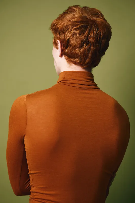 Person with curly red hair and fair skin wearing brown turtleneck sweater standing with back to camera against green background with soft lighting and shadow.