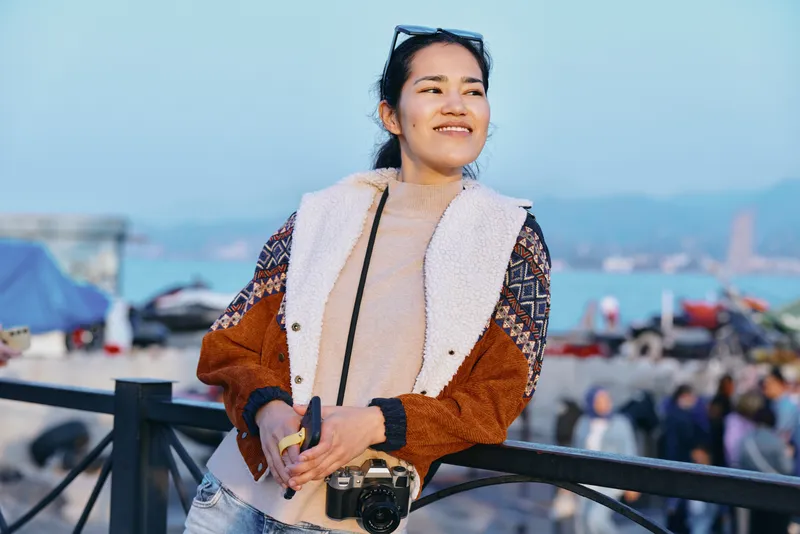 Seaside overlook with a smiling woman holding a camera, wearing a cozy jacket, candid lifestyle shot for travel, outdoors, photography, coastline, ocean breeze, boardwalk vibes, vacation mood, fresh