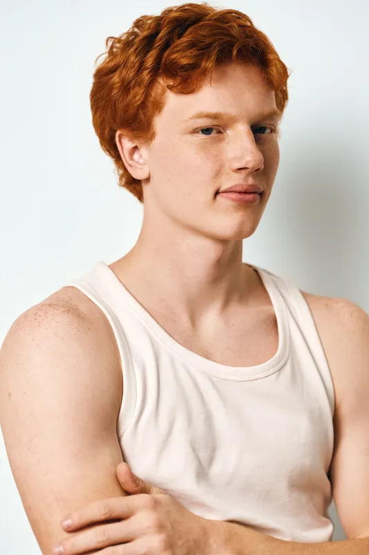 Young man with red hair wearing white tank top posing with crossed arms and calm expression against light background in studio portrait.