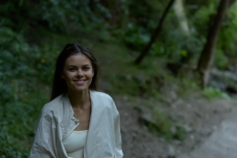 smiling woman in white jacket enjoying peaceful nature outdoors, surrounded by green forest and natural light, casual style with long dark hair, youthful expression