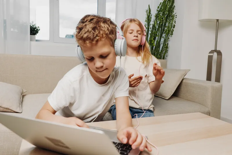 Two children enjoy technology at home, a boy works on a laptop while a girl listens to music with headphones, relaxing on a couch with a bright window background.
