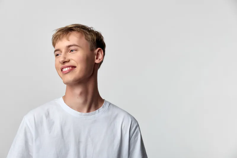Man with a bright smile wearing a white shirt stands in a clean studio, relaxed and confident, gazing off to the side, showcasing casual style and approachable mood for lifestyle photography.