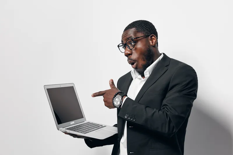Black man with dark skin wearing glasses and suit holding laptop with surprised expression on face against white background in studio.