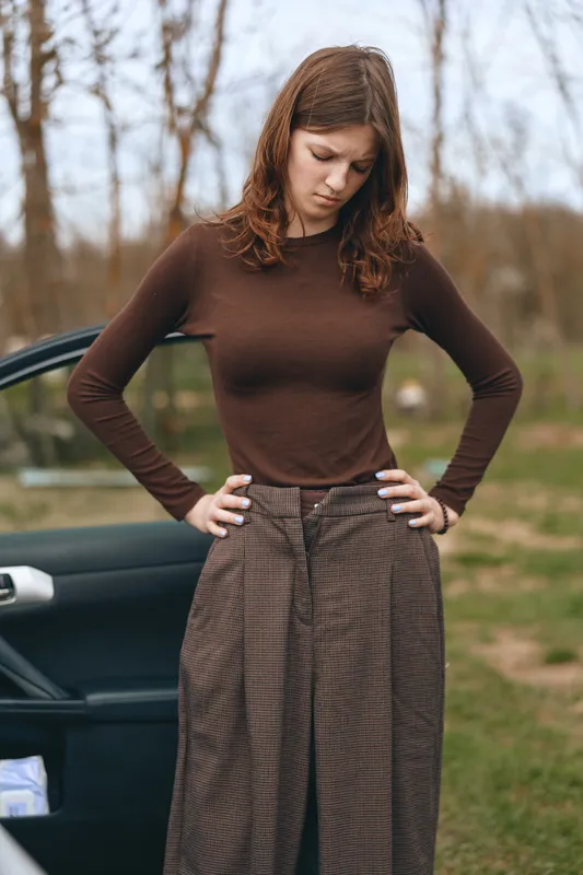 A contemplative woman stands outdoors beside an open car door, wearing a brown sheer top and checkered skirt, showcasing casual fashion and lifestyle in a rural setting.