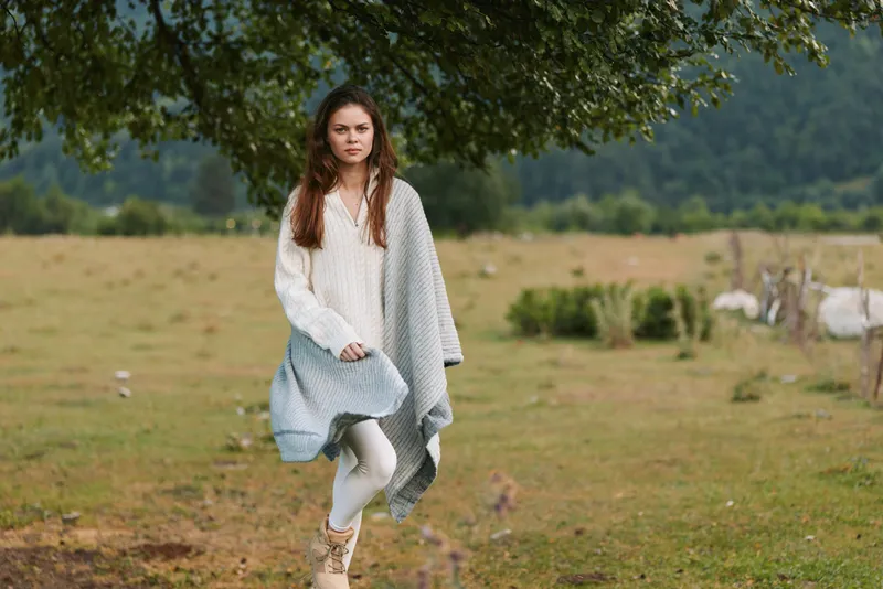 Young woman walking across a grassy field wearing a cozy shawl, natural light enhances outdoors lifestyle, casual fashion, and a carefree mood.