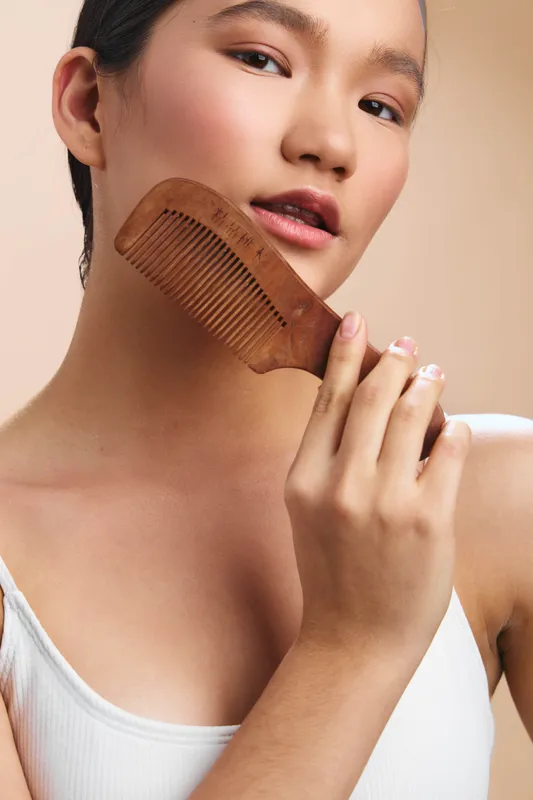 asian woman gently styles her hair with a wooden comb, showcasing skincare and beauty routine in a clean, minimal setting, conveying calm confidence and personal care.