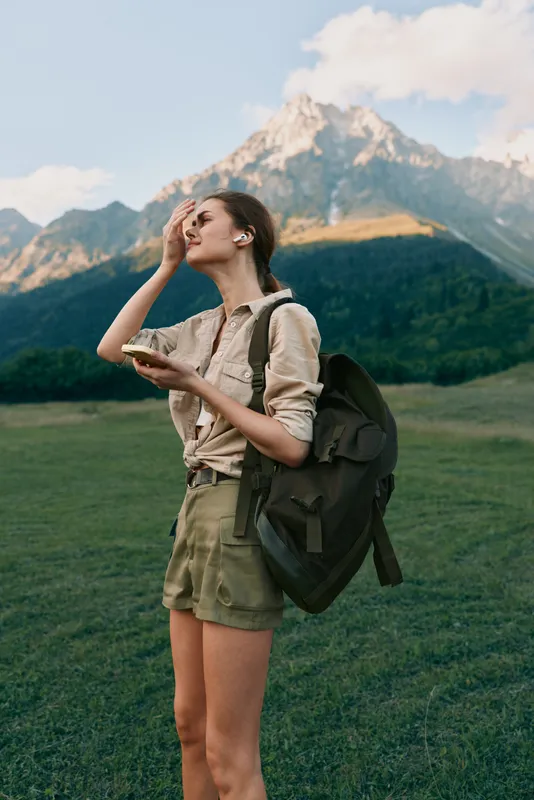 A young woman with a backpack enjoys a snack in a grassy valley with snowcapped mountains behind, capturing a moment of outdoor exploration and independence.
