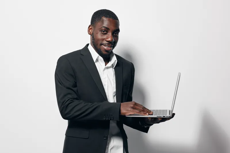 African American man with dark skin wearing black suit and white shirt using laptop computer standing against white background. Business professional working on technology with confident smile.