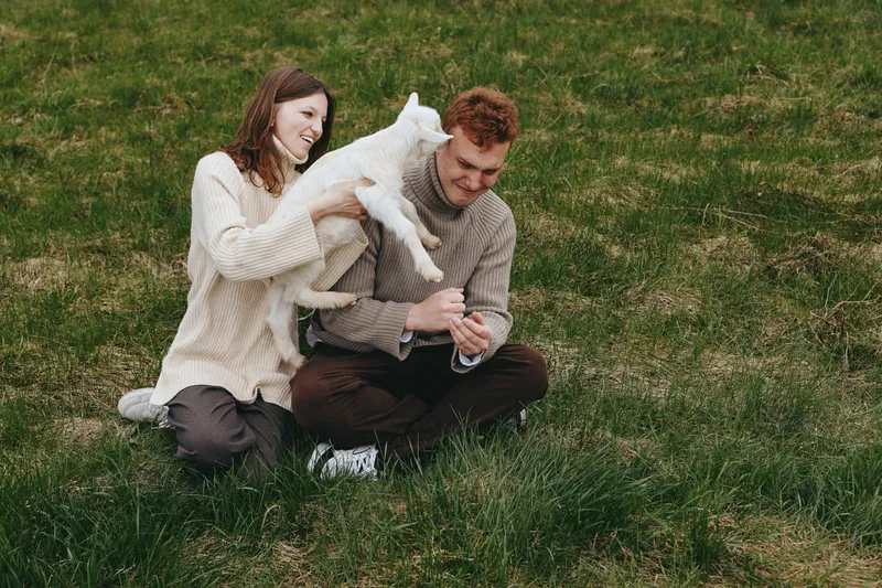 Cheerful couple outdoors, a white goat kid leaping toward them, gentle interaction, relaxed posture, sunlit grass, natural colors, candid moment, warm mood, rustic setting