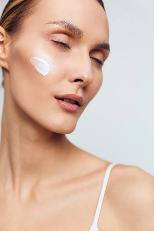 Young woman with fair skin and blonde hair applies white face cream on her cheek with closed eyes in a bright studio setting, expressing calmness and self-care.