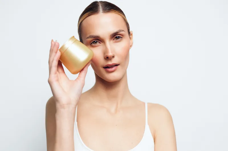 Young woman with clear skin holding a cosmetic cream jar near her face, neutral background, natural makeup, skincare concept, beauty routine, healthy lifestyle.